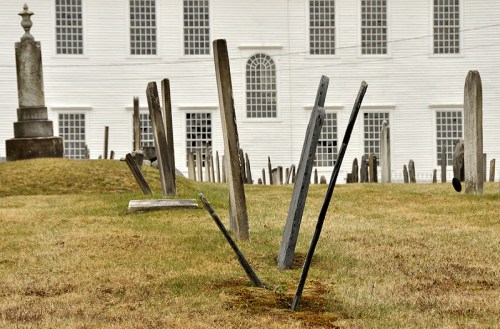 A graveyard with leaning wooden headstones on a grassy area, set against a white building in the background.