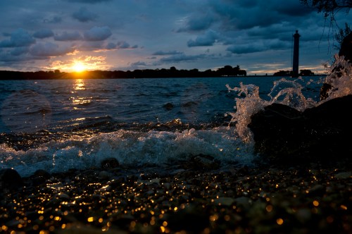 Sunset over a lake with waves crashing on the shore, a lighthouse visible in the distance against a cloudy sky.
