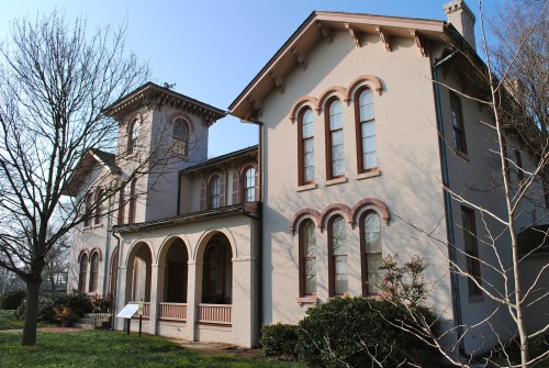 Historic two-story house with arched windows and a tower, surrounded by trees and greenery under a clear blue sky.