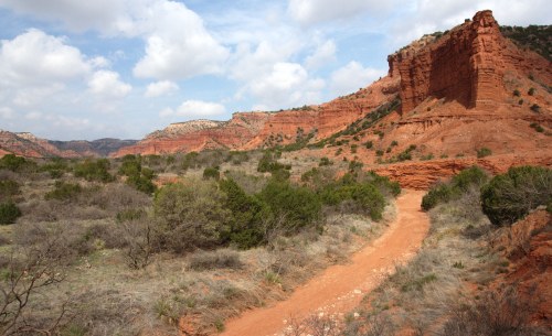 A winding dirt path leads through a rugged landscape of red rock formations and sparse vegetation under a cloudy sky.