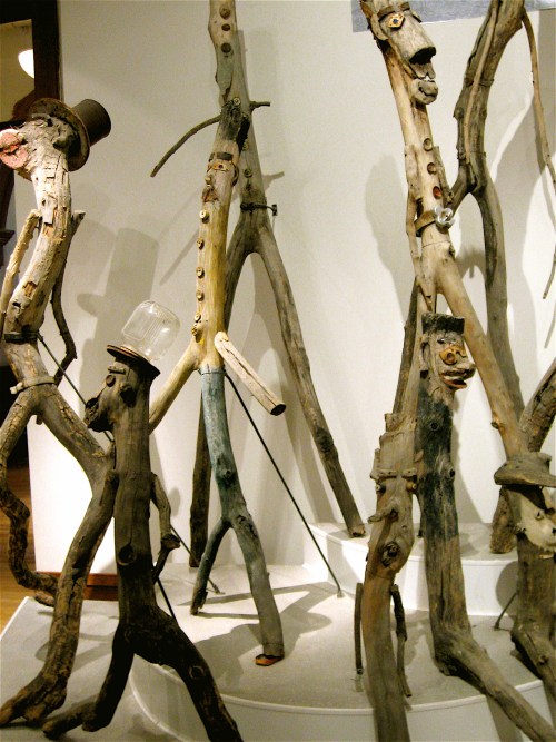 A collection of whimsical wooden sculptures resembling human figures, each with unique facial features and accessories.