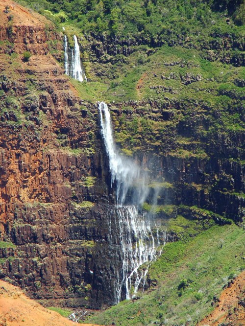 A stunning waterfall cascading down a rocky, green hillside with vibrant red earth in the foreground.