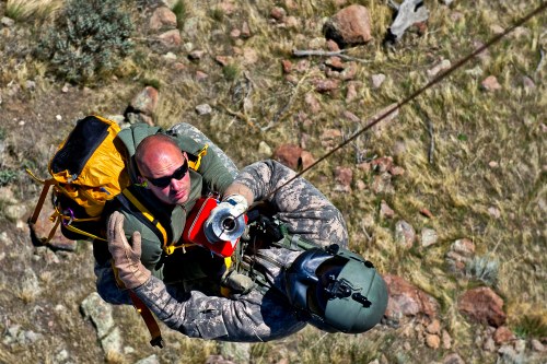 A soldier is being hoisted up by a helicopter rescue team, surrounded by rocky terrain and vegetation.