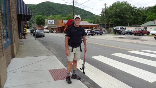 A hiker with a backpack stands on a sidewalk near a street, ready to cross in a small town surrounded by hills.
