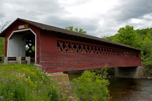 A red covered bridge spans a river, surrounded by green trees and wildflowers under a cloudy sky.