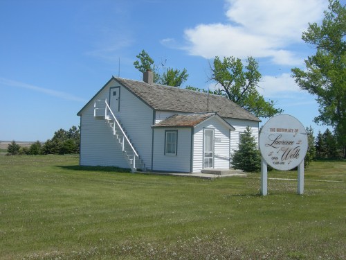 A small white house with a staircase, surrounded by green grass and trees, with a sign reading "The Birthplace of Laura Ingalls Wilder."