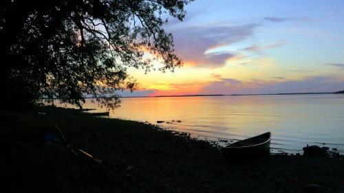 A serene lakeside scene at sunset, with a canoe on the shore and colorful clouds reflecting on the water.