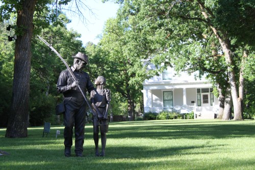 A statue of a man and a child holding a fishing rod, set in a green yard with a white house in the background.