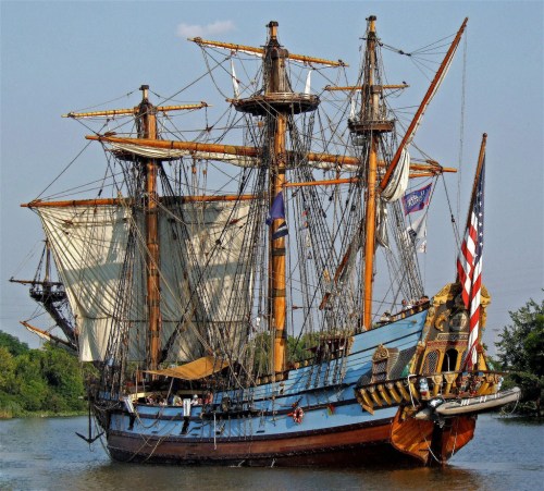 A historic tall ship with multiple masts and sails, docked on calm waters, featuring flags and a wooden hull.