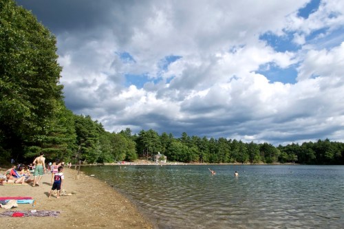 A sandy beach by a lake with people swimming and relaxing under a cloudy sky, surrounded by trees.