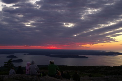 A group of people sitting on a mountain watching a colorful sunset over a calm body of water and distant islands.