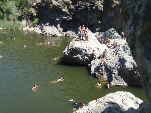 A group of people swimming and relaxing on rocks by a greenish water body surrounded by rocky cliffs and vegetation.