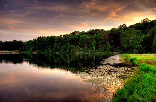 A serene lake at sunset, surrounded by lush trees and reflecting colorful clouds in the water.