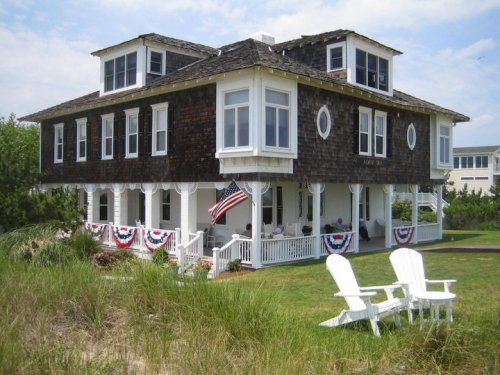 A large, two-story beach house with a shingled exterior, adorned with patriotic bunting and an American flag.