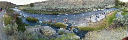 A panoramic view of a river with people enjoying the water, surrounded by rocky banks and lush greenery.