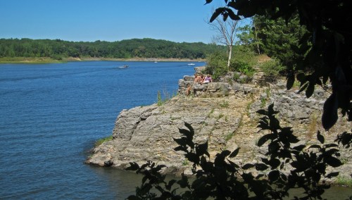 A rocky shoreline by a calm river, surrounded by greenery and trees, with people relaxing on the rocks.