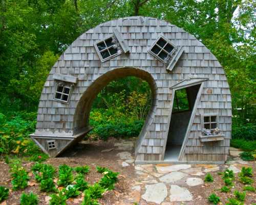 A whimsical wooden structure with a curved design and tilted windows, surrounded by greenery and flower beds.