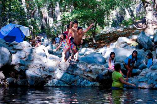 Children playing by a rocky riverbank, some jumping into the water while others relax nearby. Tents are visible in the background.