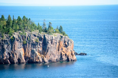 A rocky cliff covered in greenery overlooks a calm blue sea under a clear sky.