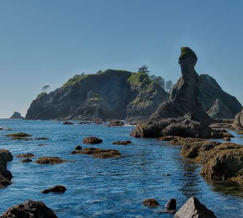 Rocky coastline with unique rock formations and lush greenery under a clear blue sky. Calm waters in the foreground.