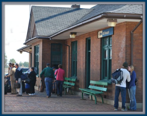 People waiting at a train station, some sitting on green benches, with a brick building in the background.