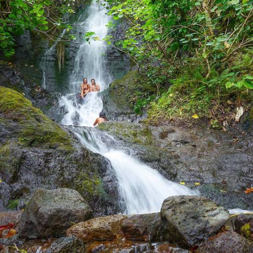 Two people sit on rocks near a waterfall surrounded by lush greenery. Water cascades down the rocky surface.