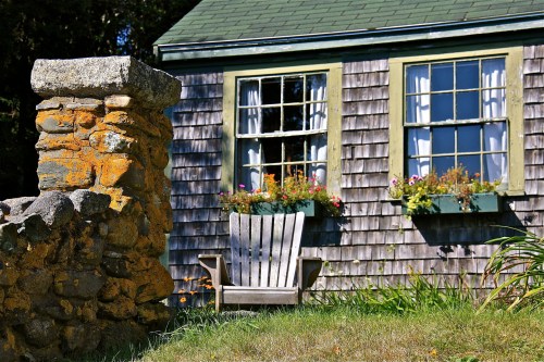 A rustic house with wooden shingles, two windows with flower boxes, and an Adirondack chair in the foreground.