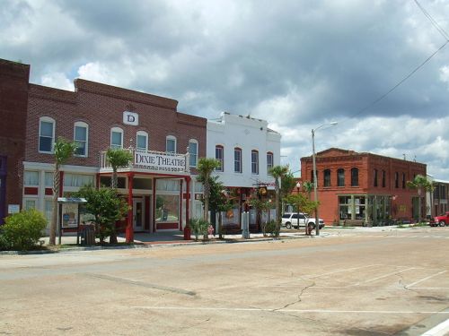 A street view of Dixie Theatre and nearby buildings under a cloudy sky, with palm trees lining the sidewalk.