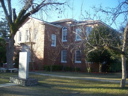 A brick building with large windows, surrounded by bare trees and a grassy area, featuring a stone monument in front.