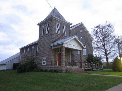 A stone church with a tall steeple, red door, and grassy surroundings under a cloudy sky.