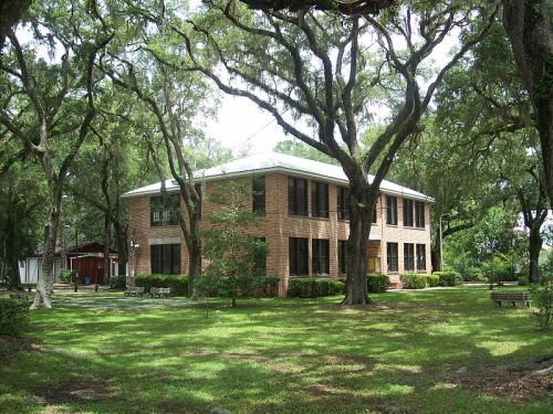 A two-story brick building surrounded by lush green trees and grass, with a bench in the foreground.