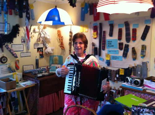 A woman in a colorful outfit plays an accordion in a cozy, eclectic room filled with various decorations and items.