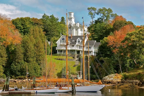A scenic view of a white mansion on a hill, surrounded by autumn trees, with sailboats in the foreground.