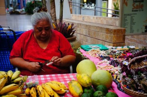 A woman sits at a table adorned with colorful crafts and fruits, focused on her work in a vibrant market setting.