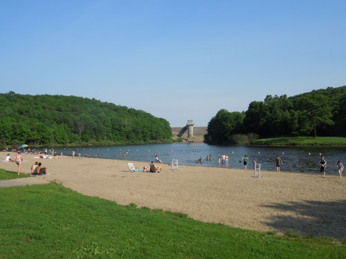 A sandy beach by a calm lake, with people swimming and enjoying the sun, surrounded by green trees and a dam in the background.