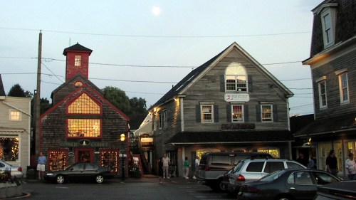 A quaint town square at dusk, featuring charming buildings, a full moon, and people strolling around.