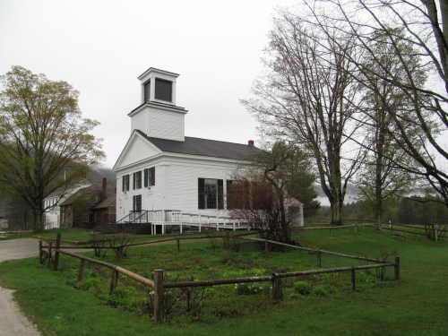 A white historic building with a bell tower, surrounded by trees and a grassy area, on a cloudy day.
