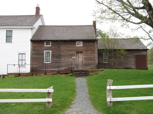 A rustic wooden house with a sloped roof, surrounded by green grass and a gravel path, next to a white building.