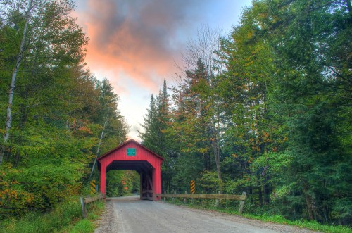 A red covered bridge surrounded by lush green trees and a colorful sky at sunset.