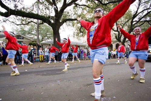A group of men in red jackets and blue shorts dance in a parade, surrounded by spectators and festive decorations.