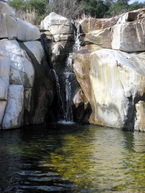 A small waterfall cascades over rocky cliffs into a clear pool of water below, surrounded by natural vegetation.