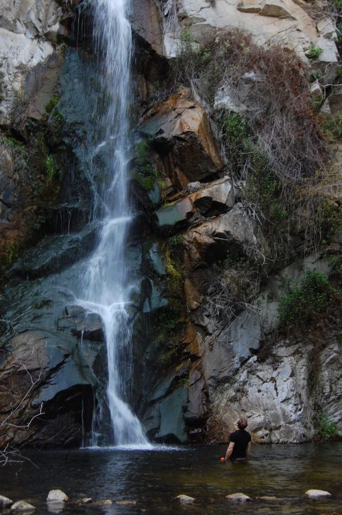 A person stands in a pool at the base of a tall waterfall, surrounded by rocky cliffs and lush greenery.