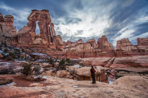 A person stands on a rocky landscape, gazing at towering rock formations under a dramatic sky.