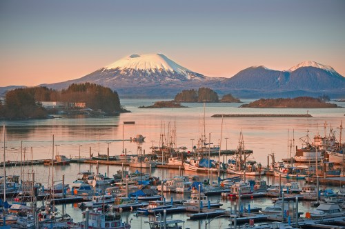 A scenic view of a marina at sunset, with boats and snow-capped mountains in the background.