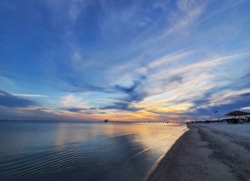 A serene beach at sunset, with calm waters reflecting colorful clouds and a pier in the distance.