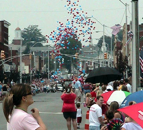 A festive street scene with a crowd watching balloons in red, white, and blue being released into the sky.