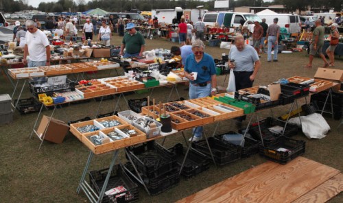 A bustling outdoor flea market with tables of various tools and items, and shoppers browsing among the stalls.