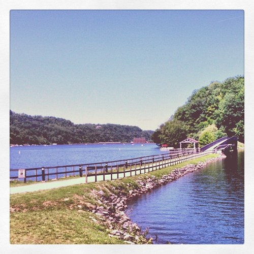 A serene river scene with a path along the water, surrounded by lush green hills under a clear blue sky.