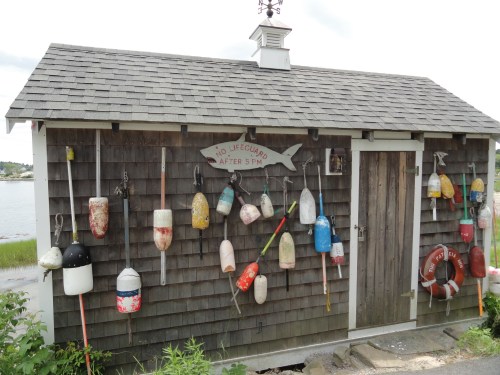 A rustic shed with a shingled roof, adorned with colorful buoys and a lifebuoy, near a body of water.