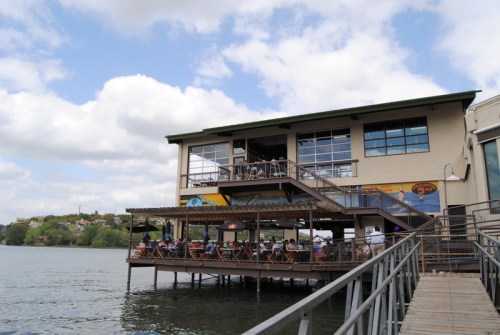 A waterfront restaurant with outdoor seating, overlooking a lake, under a partly cloudy sky.